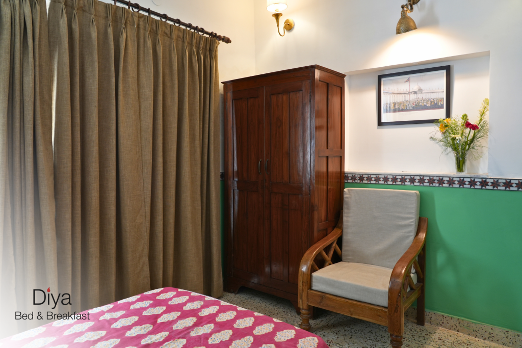 Cozy bedroom corner at Diya Bed & Breakfast featuring a wooden wardrobe, cushioned armchair, wall art, and fresh flowers in a vase.