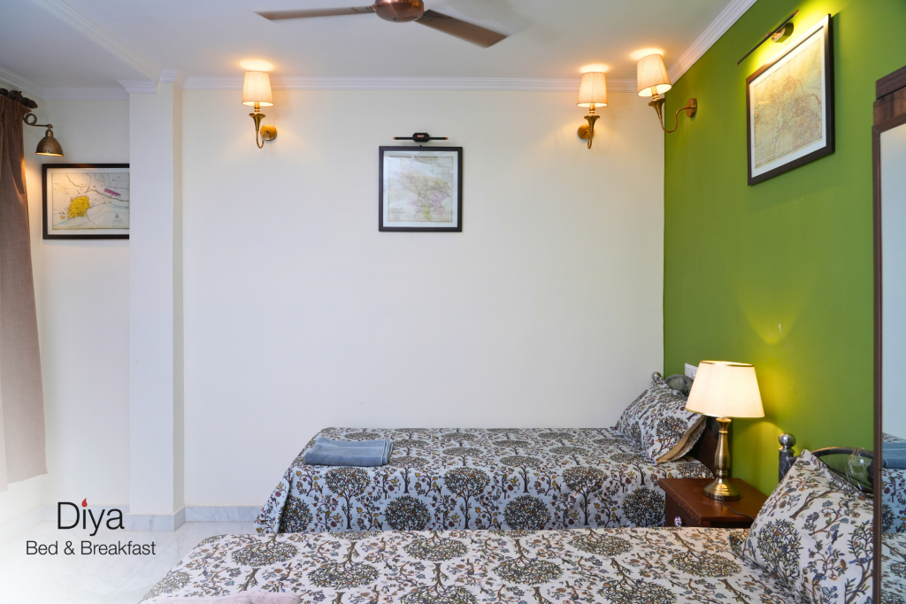 A cozy twin-bed room at Diya Bed & Breakfast with patterned bedspreads, green accent wall, wall-mounted lamps, and framed maps.