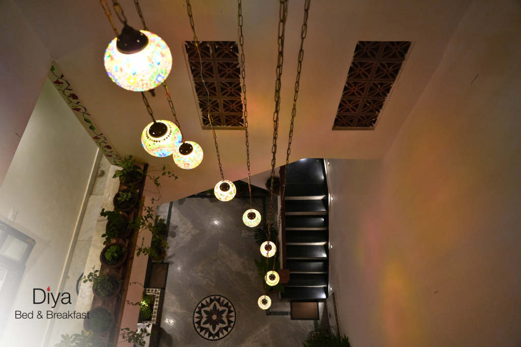 Hanging mosaic pendant lights illuminating a staircase area at Diya Bed & Breakfast, with indoor plants and patterned marble flooring.