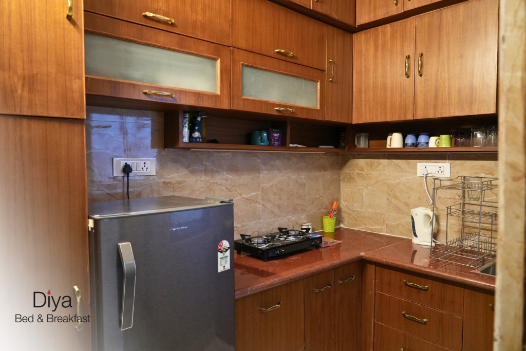 Compact kitchen at Diya Bed & Breakfast with wooden cabinets, a small refrigerator, countertop gas stove, electric kettle, dish rack, and neatly arranged cups and glasses.