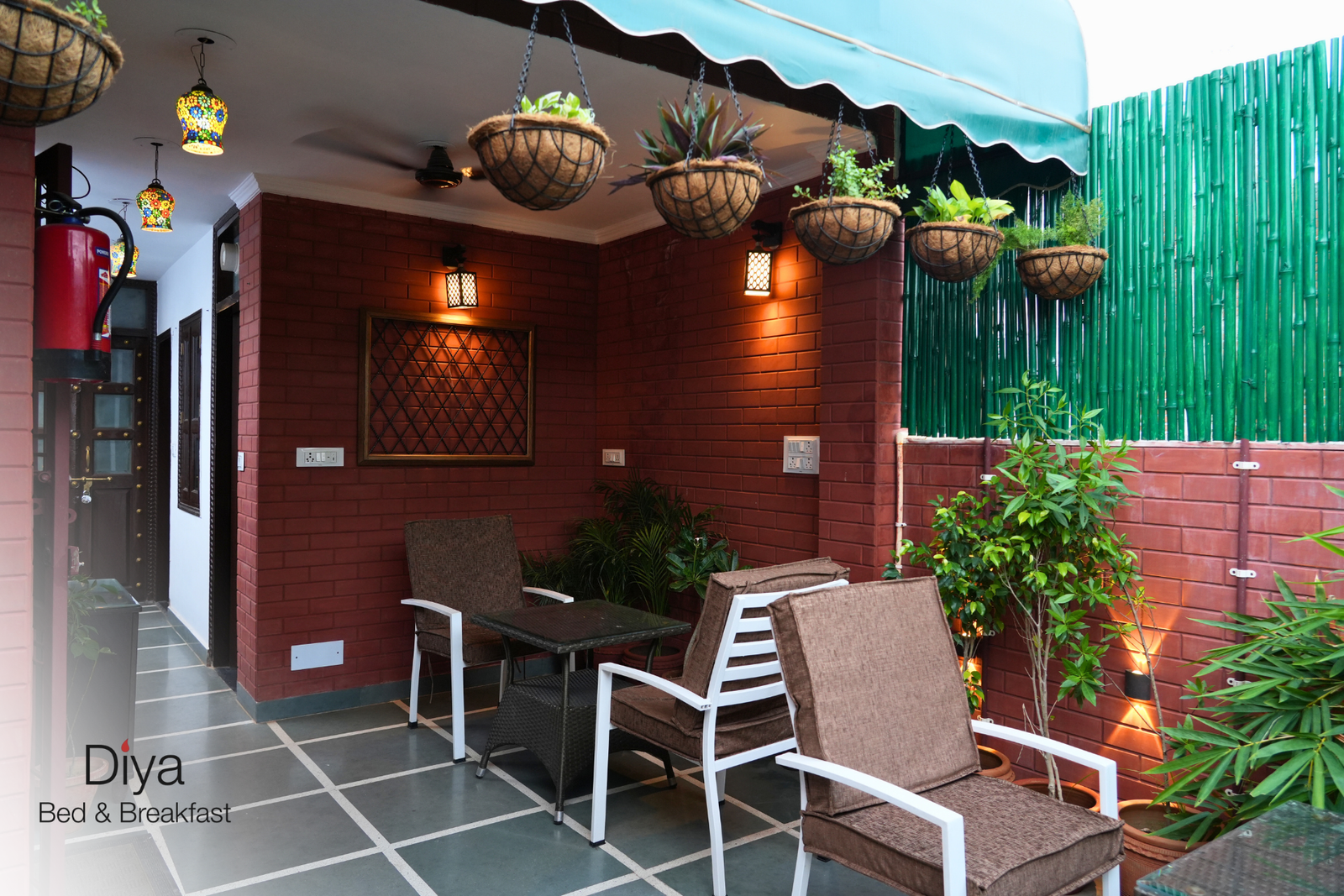 Outdoor seating area of Diya Bed & Breakfast with brown wicker chairs, a small glass-top table, hanging planters, green bamboo fencing, and warm wall lighting.