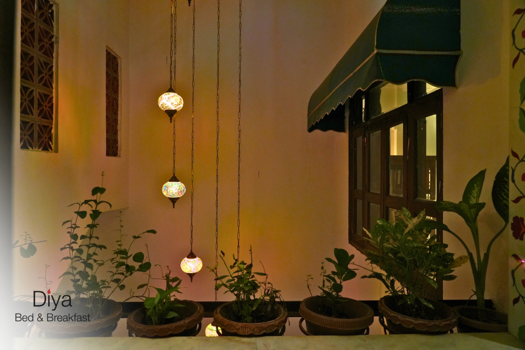 Colorful mosaic glass pendant lamps hanging vertically beside potted plants at Diya Bed & Breakfast, with warm lighting and a green awning over a wooden window.