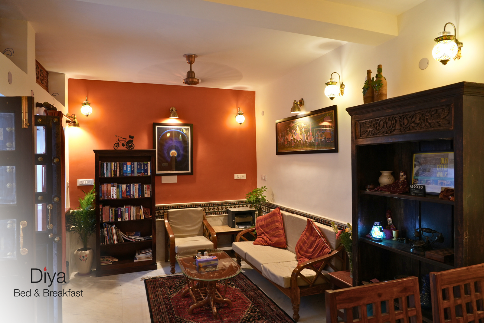 Cozy lounge area at Diya Bed & Breakfast with warm lighting, bookshelf, wooden furniture, red accent wall, and decorative art pieces.