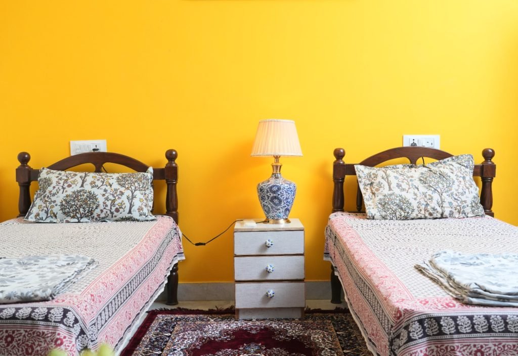 Bedroom with two single beds, patterned bedding, a shared nightstand, and a blue decorative lamp against a bright yellow wall.