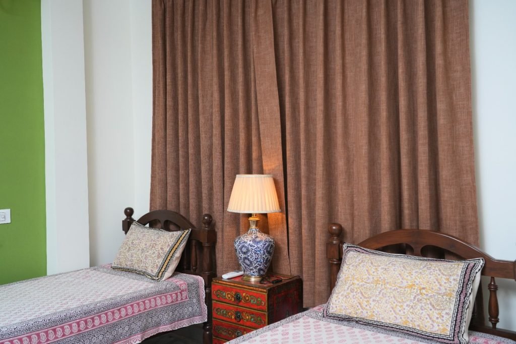 Two single beds with patterned bedspreads and pillows, separated by a decorative lamp on a red side table, in front of closed brown curtains.