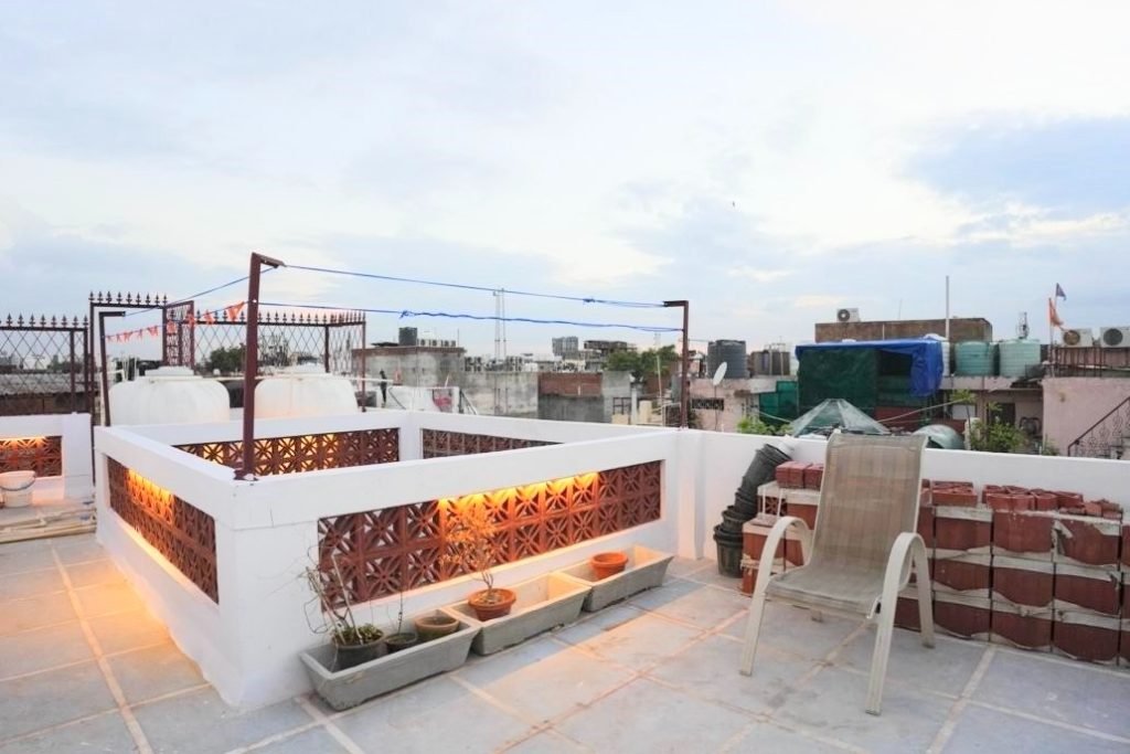 Rooftop terrace with potted plants, a plastic chair, and surrounding cityscape under a cloudy evening sky.
