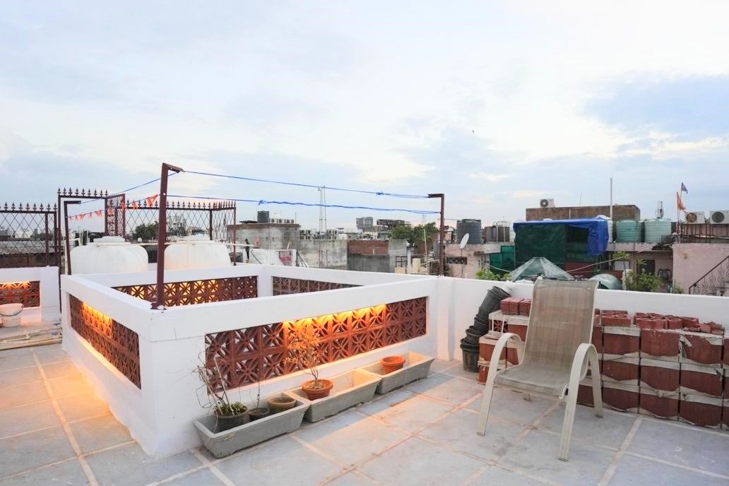 Rooftop terrace with potted plants, a plastic chair, and surrounding cityscape under a cloudy evening sky.