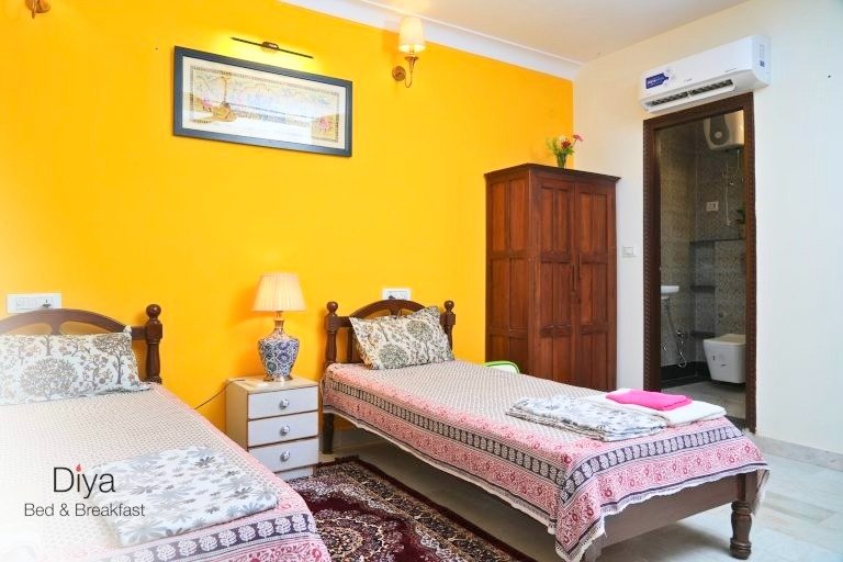 Bright twin bedroom at Diya Bed & Breakfast with yellow accent wall, patterned bedspreads, bedside table with lamp, wooden wardrobe, and attached bathroom.