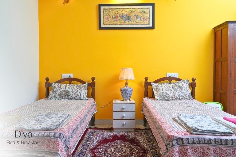 Two single beds with patterned bedding, a central nightstand with a blue and white ceramic lamp, a yellow accent wall, and a framed traditional painting above.