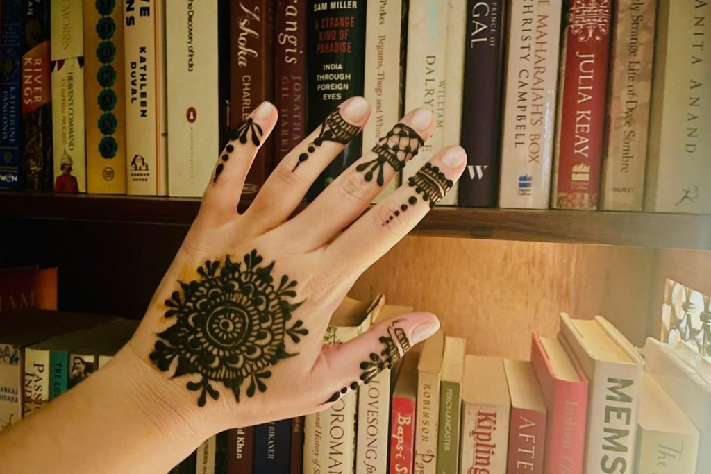 Hand decorated with intricate mehndi design, held up in front of a bookshelf filled with various books.
