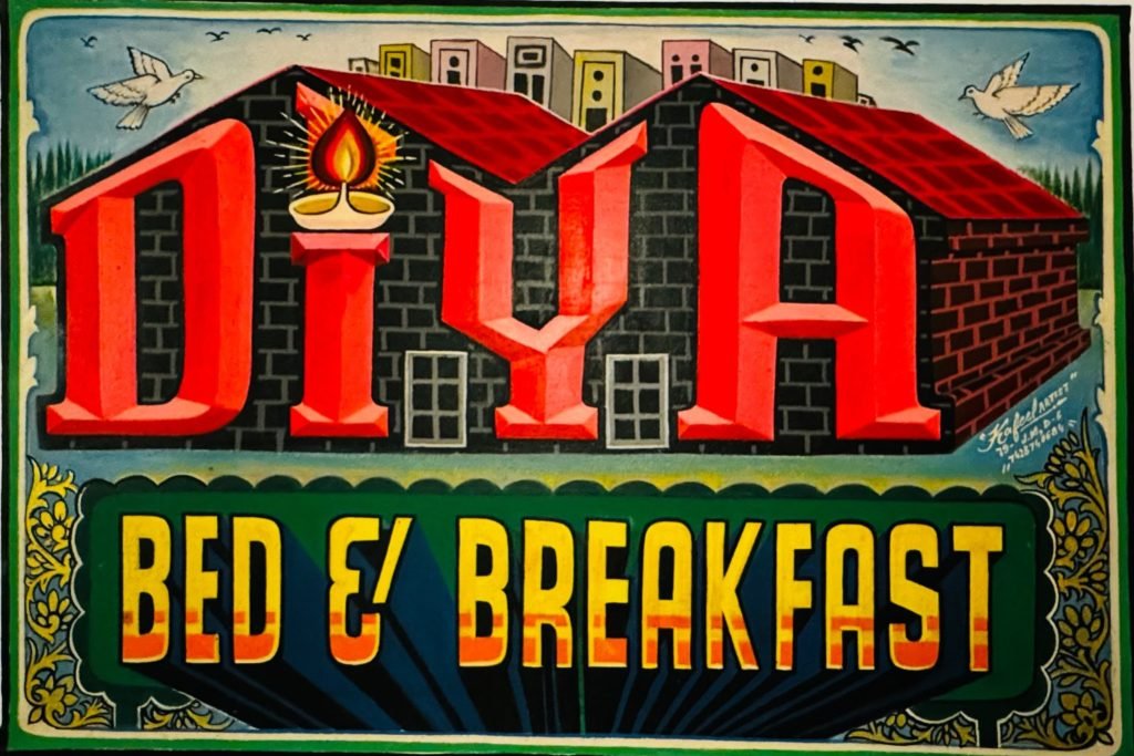 Colorful hand-painted sign for "Diya Bed & Breakfast" with bold red and yellow letters, featuring a traditional oil lamp and building illustration.
